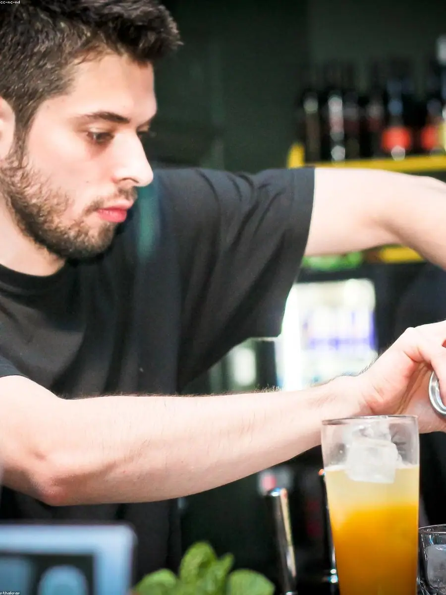 Bartender pouring a cocktail in a long stream behind a dim, moody bar